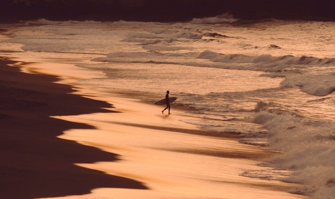 Surfer walking along a reflective beach beside breaking waves