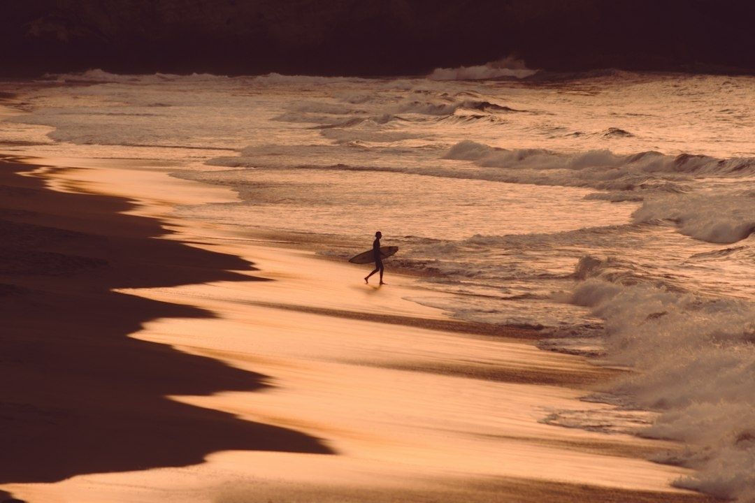 Surfer walking along a reflective beach beside breaking waves